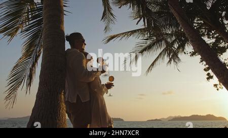 SILHOUETTE ORO SCURO VISTA TRAMONTO: Coppia amorevole in camicia bianca e occhiali da sole, vicino alla palma, baciare, abbracciare, rilassarsi e bere cocktail. Concetto tropicale vacanze estive, vacanze matrimonio amore Foto Stock