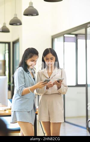 Due dipendenti donne che cercano uno smartphone e parlano con il partner mentre si trovano in un ufficio moderno. Colleghi che hanno un chill parlare in ufficio. Foto Stock