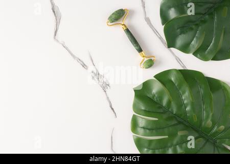 Rullo facciale in pietra di Giada con foglie verdi di monstera su sfondo di marmo bianco. Massaggiatore per il viso. Disposizione piatta. Spazio di copia Foto Stock