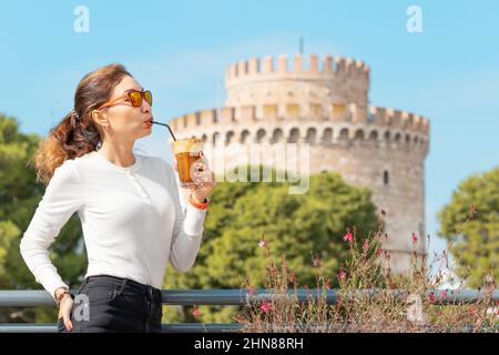 Una ragazza sorridente beve una bevanda dolce di caffè Frappe, inventata a Salonicco, Grecia. Sullo sfondo della famosa Torre Bianca. Foto Stock