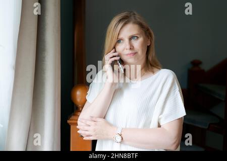 Donna adulta felice che parla al telefono mentre si alza alla finestra contro le scale di sfondo nel soggiorno. La donna caucasica comunica utilizzando la connessione cellulare in abiti casual, guarda verso la finestra Foto Stock