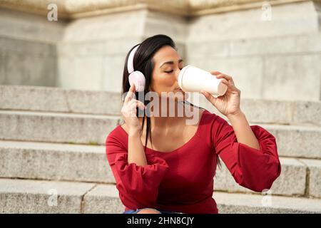 Attraente femmina ispanica che ascolta la musica in cuffie wireless mentre beve gustosa bevanda calda da asporto su scale di pietra in città Foto Stock