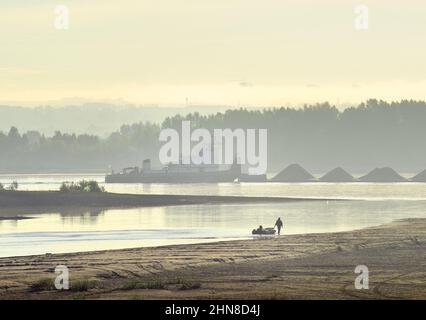 Novosibirsk regione, Siberia, Russia - 08.20.2020: Un pescatore cammina lungo la riva del fiume OB. Una chiatta di carico naviga attraverso la nebbia mattutina Foto Stock