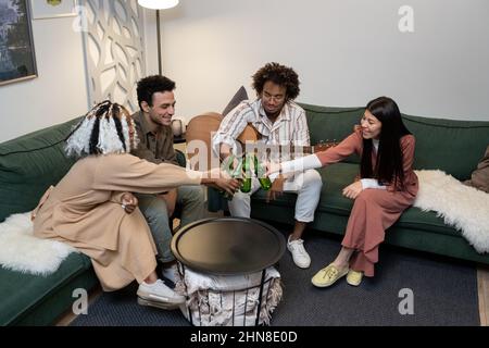 Gruppo di amici felici che tostano con bottiglie di birra mentre si siedono sul divano e suonano la chitarra durante le feste a casa Foto Stock