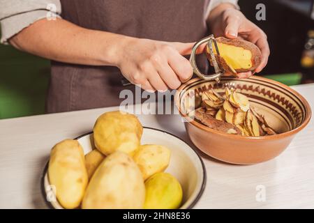 le mani di cuoco tagliano la buccia delle patate crude con il coltello da cucina per sbucciare le verdure, sbucciano, mettono i rifiuti alimentari in un contenitore separato per ulteriore lavorazione. Foto Stock