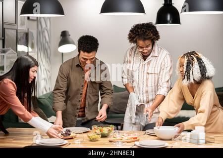 Gruppo di giovani amici che mettono insieme il tavolo da pranzo mettendo il pasto e i piatti, si preparano per la cena partito Foto Stock