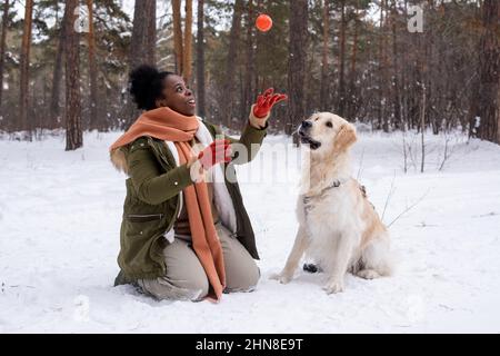 Donna africana in abiti caldi che gioca la palla con il suo cane durante la loro passeggiata nella foresta in inverno Foto Stock