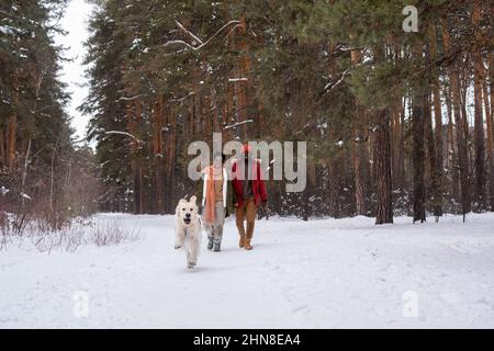 Felice coppia africana in abiti caldi che camminano lungo il percorso nella foresta invernale con il loro cane da corsa Foto Stock
