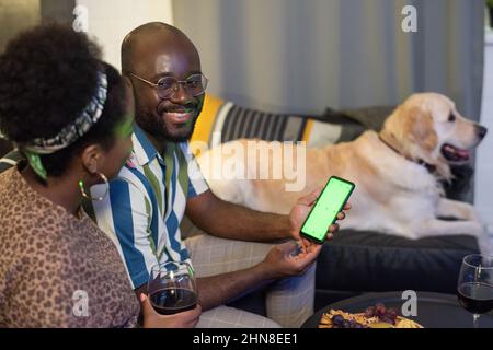 L'uomo africano guarda video divertenti sul telefono cellulare insieme alla donna mentre si siedono sul divano in camera Foto Stock