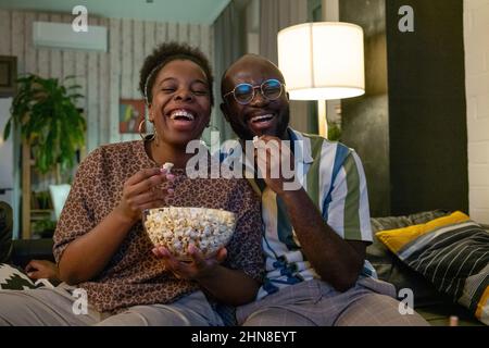 Felice coppia africana mangiare popcorn durante la commedia e ridere seduto sul divano in salotto Foto Stock