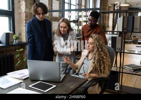 Donna d'affari incinta che punta al laptop e discute di presentazione online con i suoi colleghi durante il lavoro di squadra in ufficio Foto Stock