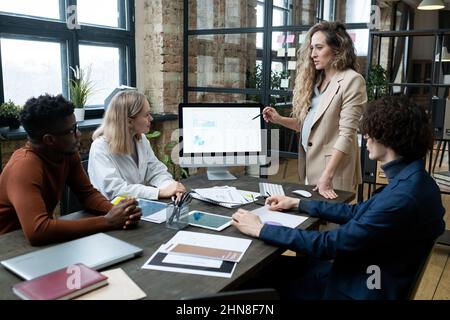 Donna d'affari incinta che punta al monitor del calcolatore con i grafici finanziari e che parla con i suoi colleghi durante la presentazione di affari all'ufficio Foto Stock