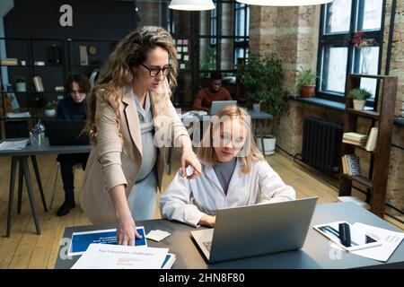Giovane donna d'affari incinta che punta al laptop e discute di lavoro in linea con il suo collega durante il lavoro in ufficio Foto Stock