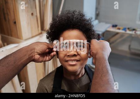 Primo piano di un irriconoscibile uomo afroamericano che dà occhiali protettivi a simpatico figlio aiutare padre in officina Foto Stock