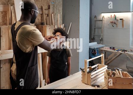 Ritratto di nero padre e figlio lavorando insieme in officina garage, concentrarsi sul ragazzo sorridente indossare occhiali di sicurezza, spazio copia Foto Stock