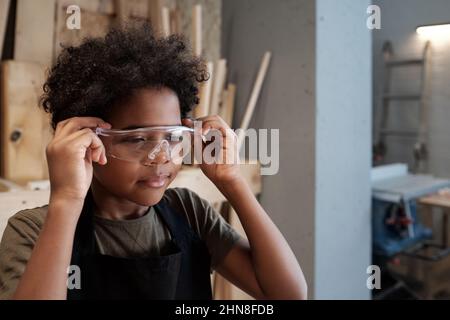 Ritratto di ragazzo afroamericano carino indossare occhiali protettivi mentre aiutare il padre in officina, copia spazio Foto Stock