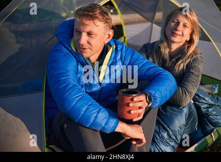 Bel giovane uomo che tiene una tazza di caffè, seduto accanto a donna affascinante in tenda turistica. Bella coppia di escursionisti che riposano nella tenda del campo. Concetto di viaggio, campeggio e relazioni. Foto Stock