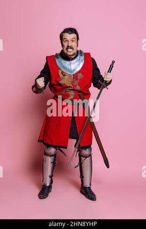 Un giovane uomo, guerriero medievale o cavaliere che indossa abiti da braccio che posano isolati su sfondo rosa. Confronto di epoche, storia, emozioni Foto Stock