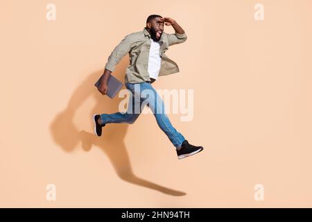 Foto a tutta lunghezza del profilo di giovane barba impressionato ragazzo run hold laptop indossare camicia jeans sneakers isolato su sfondo beige Foto Stock