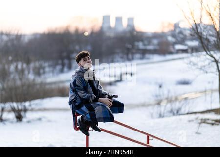 Ritratto di un giovane uomo in un cappotto di plaid e sciarpa sullo sfondo di un paesaggio industriale in inverno. Foto Stock
