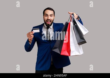 Uomo portatore che tiene e mostra borse di carta e carta di credito, pagamenti contactless, shopping facile con carta di debito, vestito di stile ufficiale. Studio interno girato isolato su sfondo grigio. Foto Stock