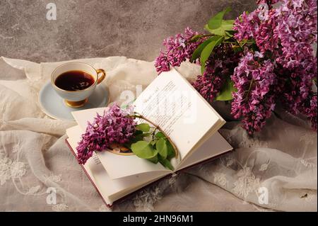 Libro di cucina, caffè e bouquet lilla disposti splendidamente su un panno da tavolo bianco ricamato. Foto Stock