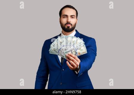 Ritratto di sicuro di sé ricco barbuto uomo che tiene fuori bollette dollaro a macchina fotografica, vantando soldi vinti in lotteria, indossando vestito di stile ufficiale. Studio interno girato isolato su sfondo grigio. Foto Stock