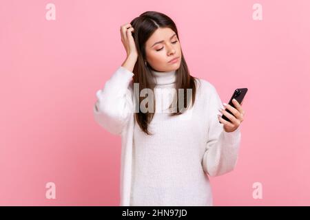 Donna giovane e pensiva che tiene in mano lo smartphone, con un'espressione facciale premurosa, un tocco di testa e un pullover casual bianco. Studio interno girato isolato su sfondo rosa. Foto Stock