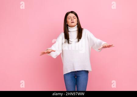 Ritratto di giovane donna disorientata che cammina con gli occhi chiusi e che tiene le mani per trovare la strada, indossando bianco stile casual maglione. Studio interno girato isolato su sfondo rosa. Foto Stock