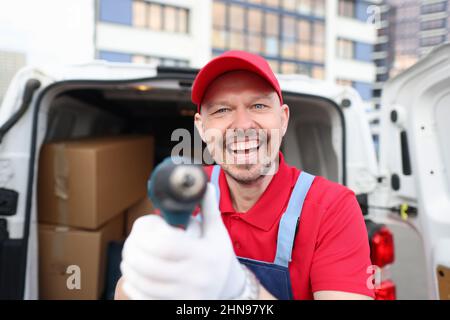 Un corriere felice in una divisa rossa mostra uno strumento vicino ad un furgone coperto Foto Stock