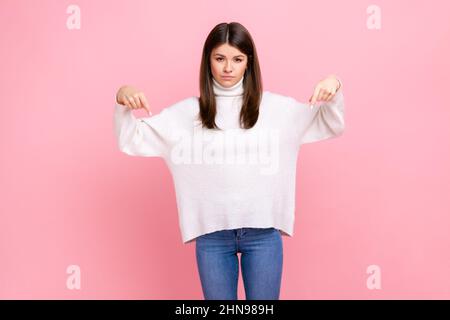 Donna puntando le dita verso il basso, impegnativo approccio a lei immediatamente, avendo il controllo sulla situazione, indossando bianco stile casual maglione. Studio interno girato isolato su sfondo rosa. Foto Stock