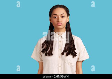 Ritratto di donna seria con dreadlocks neri, guardando attentamente la macchina fotografica, avendo l'espressione facciale severa bossy, indossando la camicia bianca. Studio interno girato isolato su sfondo blu. Foto Stock