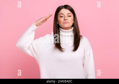 Ritratto di donna seria responsabile saluta comandante, ordine d'ascolto con espressione obbediente, indossando bianco casual stile maglione. Studio interno girato isolato su sfondo rosa. Foto Stock