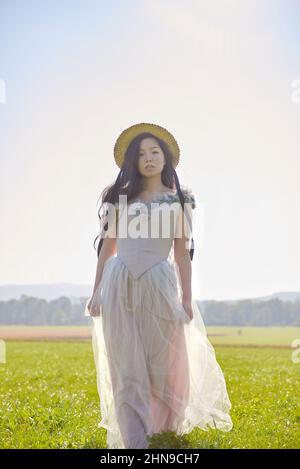 Giovane bella lunga capelli neri asiatica donna che indossa un abito romantico lilla in piedi in un campo d'erba retroilluminato Foto Stock