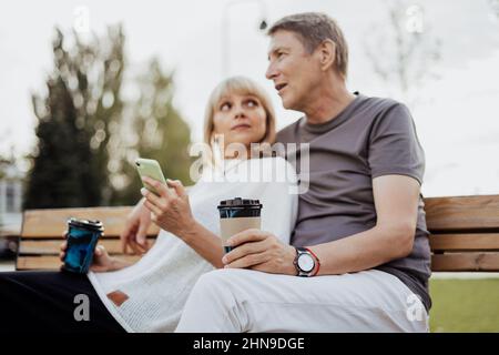Una coppia adulta felice in amore seduto su panca con telefono all'aperto nel parco. Un biondo caucasico uomo e donna passare il tempo insieme e bere Foto Stock