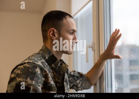 Soldato depresso e triste in uniforme verde con trauma dopo la guerra in piedi vicino alla finestra Foto Stock