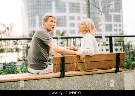Un adulto maturo felice coppia romantica in amore seduto su panchina all'aperto nel parco di strada della città. Un biondo uomo e una donna caucasica trascorrono del tempo insieme Foto Stock