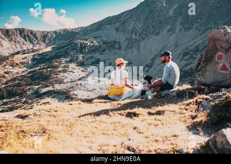 coppia con cane seduto su rocce ammirando il paesaggio di montagna Foto Stock