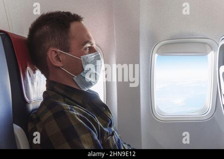 Un giovane uomo in una maschera medica blu siede nel sedile passeggero a bordo dell'aereo e guarda fuori la finestra. Volo sicuro durante lo scoppio del covi Foto Stock