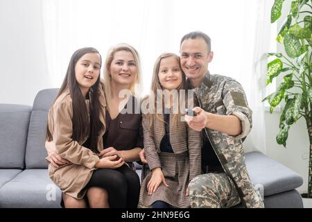 Giovane soldato coraggioso che trascorre del tempo con la sua famiglia. Stanno guardando la televisione. Uomo con telecomando in mano. Foto Stock