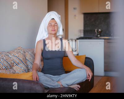 donna seduta sul divano, posizione lotus mette le mani sulle ginocchia, occhi chiusi, godere di meditazione, fare yoga. Niente stress, abitudini sane, stile di vita cosciente, concetto di sollievo dall'ansia Foto Stock