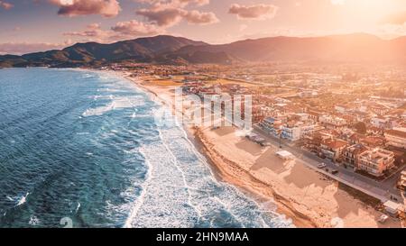 Spettacolare vista aerea panoramica dell'iconica cittadina turistica di Sarti e della famosa spiaggia di sabbia lunga e vuota al tramonto con onde alte e bagliore al sole Foto Stock