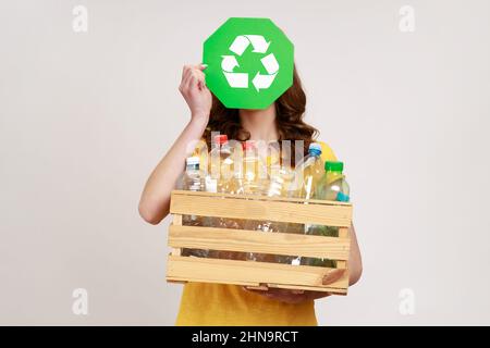 Femmina sconosciuta che tiene la scatola di legno piena di bottiglie di plastica e la faccia di copertura con canta di riciclo. Giornata ambientale e concetto di riciclaggio. Studio interno girato isolato su sfondo grigio. Foto Stock