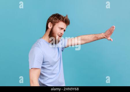 Vista laterale di un uomo stretto barbuto che mostra un segnale di stop, in piedi con il viso accigliato e l'espressione bossy, che significa cautela per evitare pericolo o errore. Studio interno girato isolato su sfondo blu. Foto Stock