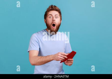 Ritratto di sorpreso giovane adulto bello ported uomo che legge la lettera o il biglietto di auguri, tenendo la busta, essendo stupito di notizie scioccate. Studio interno girato isolato su sfondo blu. Foto Stock