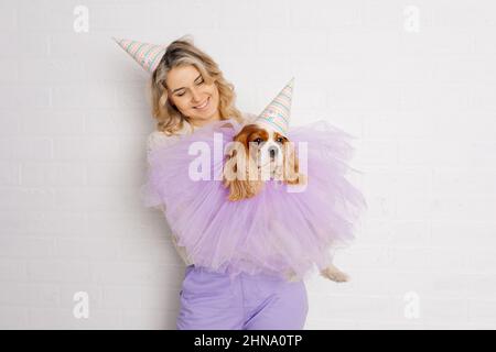 Ritratto di giovane donna che tiene il suo animale domestico Cavalier re Charles Spaniel. Sono vestiti in bianco e lilla colori e identici cappelli divertenti per le feste Foto Stock