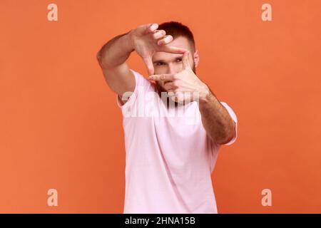 Ritratto di concentrato creativo bearded uomo alzando le mani agli occhi e facendo cornice alla ricerca di un colpo adatto, cameraman lavoro, indossando T-shirt rosa. Studio interno girato isolato su sfondo arancione. Foto Stock