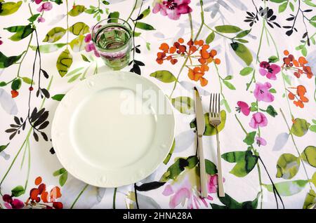 set da tavolo con tovaglia floreale, piatto bianco, vetro verde e posate in acciaio inox Foto Stock