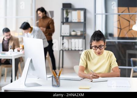 Giovane donna d'affari in occhiali seduti sul posto di lavoro con il computer e prendere appunti in notebook durante il suo lavoro in ufficio Foto Stock
