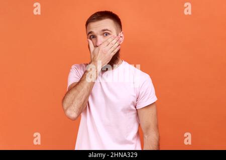 Ritratto di uomo bearded che copre la bocca con la mano per tenersi in silenzio, paura di dire segreto, guardando con espressione intimidita, indossando la T-shirt rosa. Studio interno girato isolato su sfondo arancione. Foto Stock